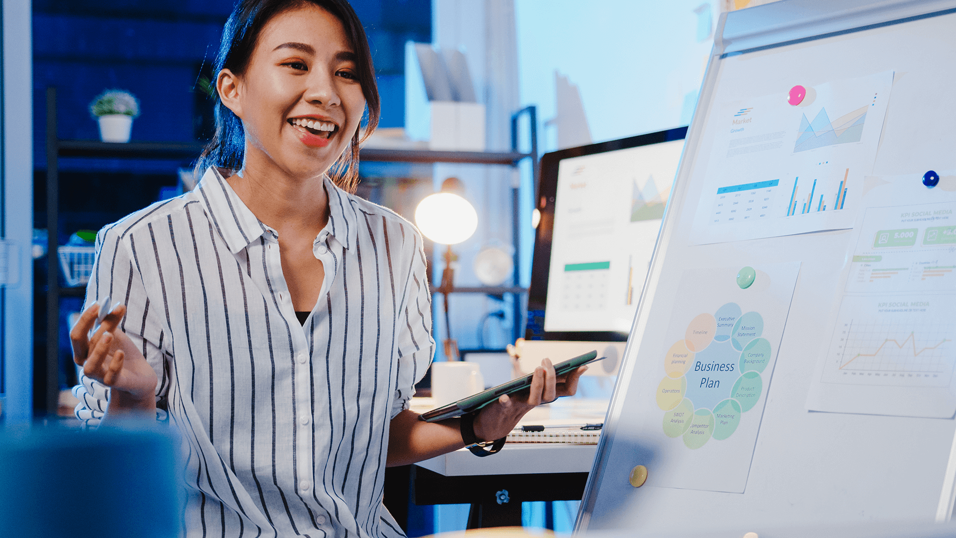 A confident professional woman presenting a business plan with colorful charts and data on a whiteboard in a modern office setting at night.