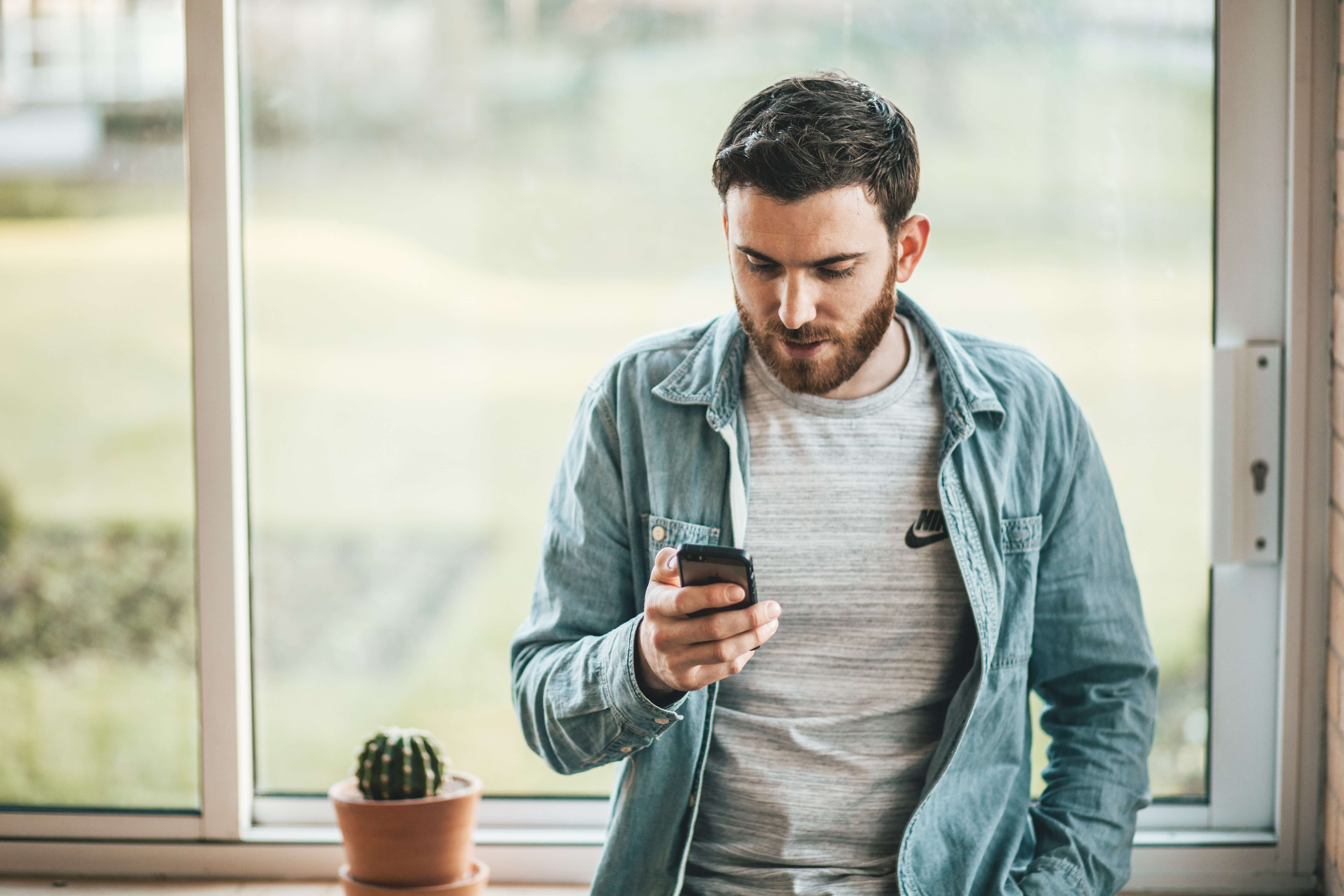 A focused young man in a casual denim jacket and grey shirt intently checking his smartphone by a bright window, with a small potted cactus on the windowsill adding a touch of greenery to the serene indoor setting.