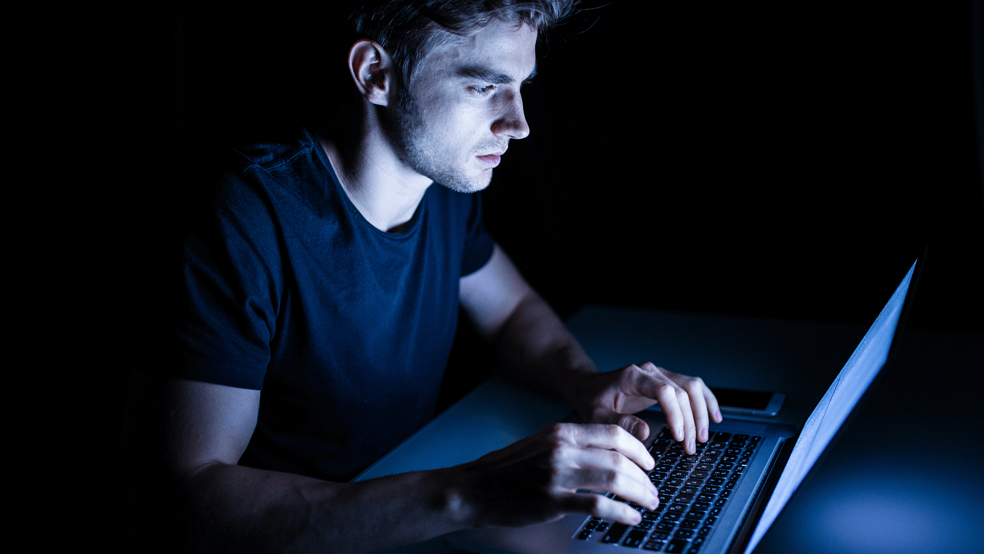 A man focused on his laptop screen in a dimly lit room, illuminated only by the blue light of the computer display.
