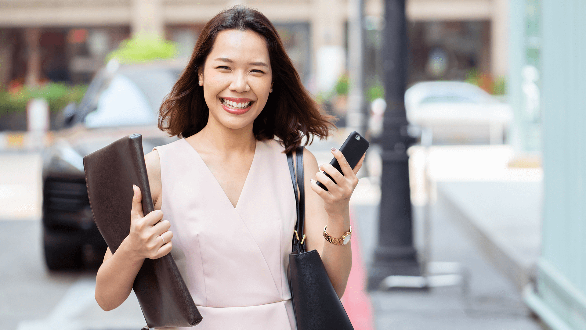 A cheerful woman in business casual attire holding a smartphone and a clutch, walking down the street.