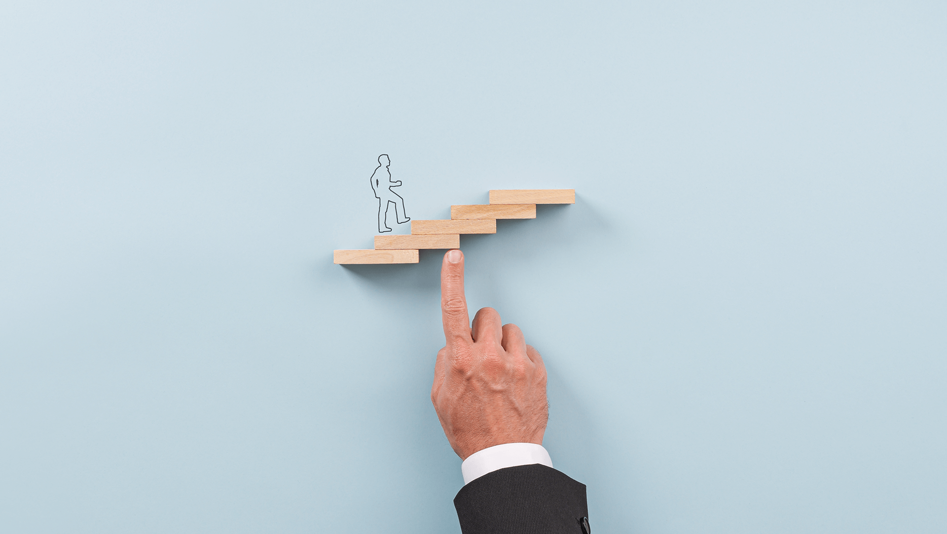 A hand boosts a paper cutout of a person up a staircase of wooden blocks against a blue background, symbolizing support and progression.