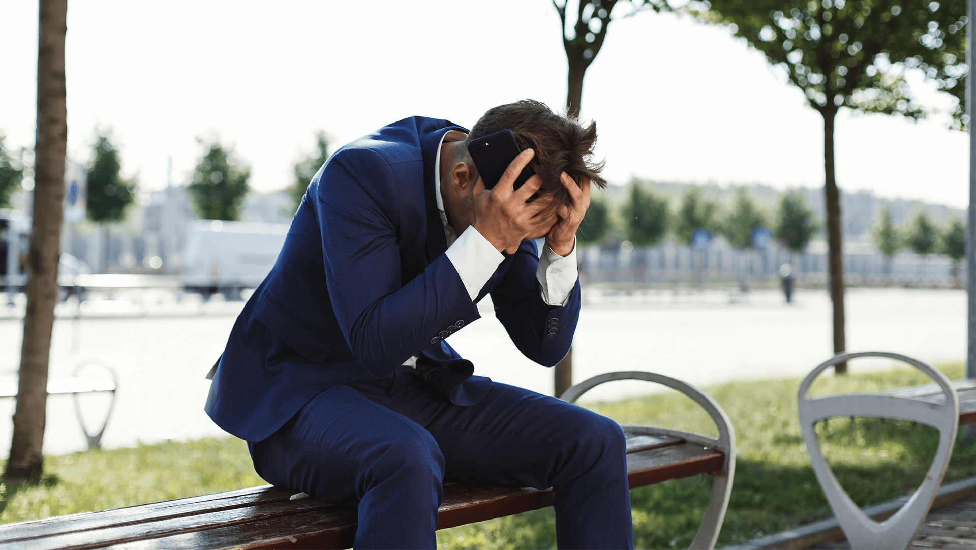 A businessman in a blue suit sitting on a park bench, looking stressed or upset while talking on his cellphone.