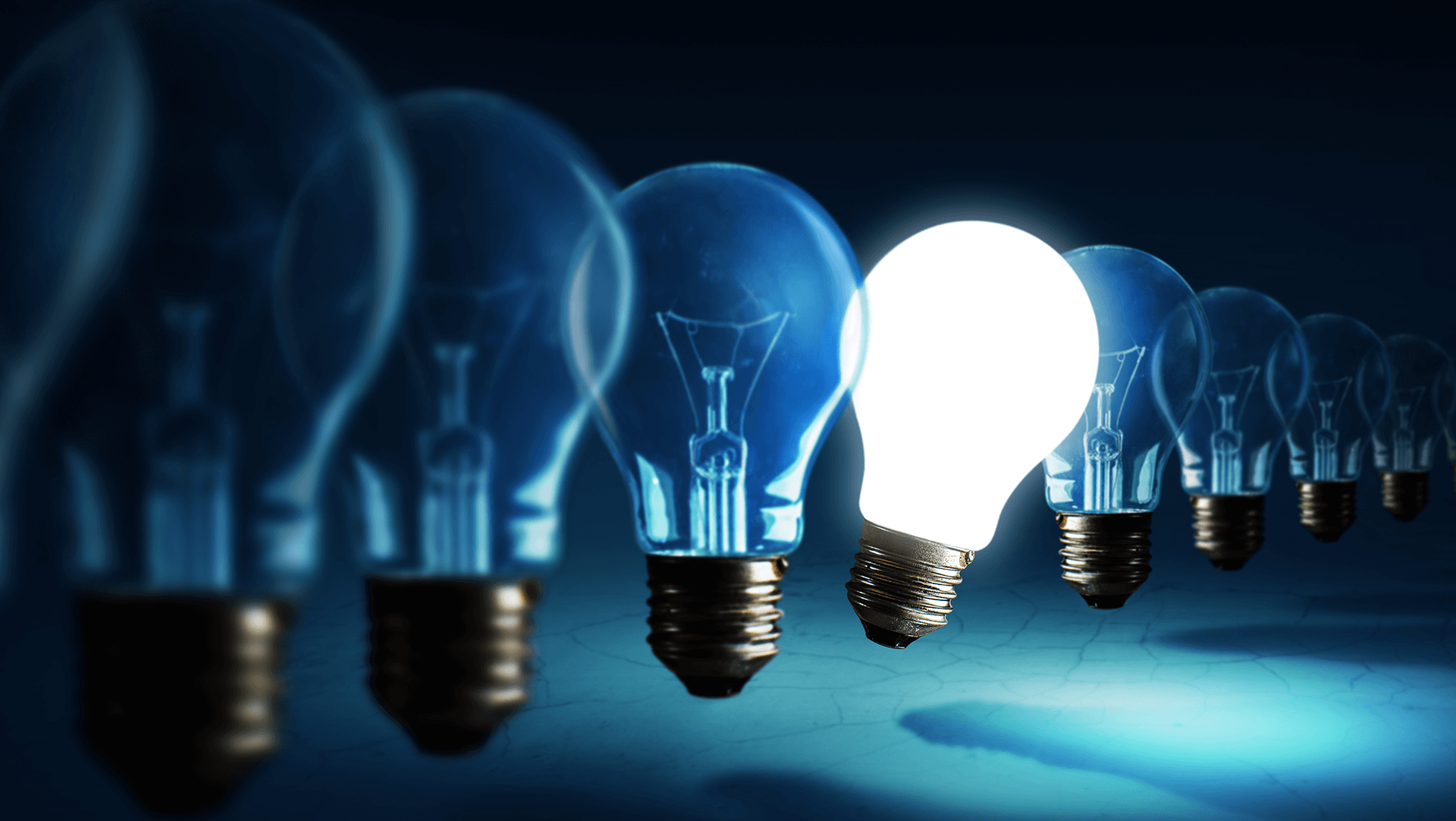 A glowing idea stands out among dim ones – an illuminated light bulb shines brightly in a lineup of unlit bulbs against a dark blue background, symbolizing inspiration and innovation.