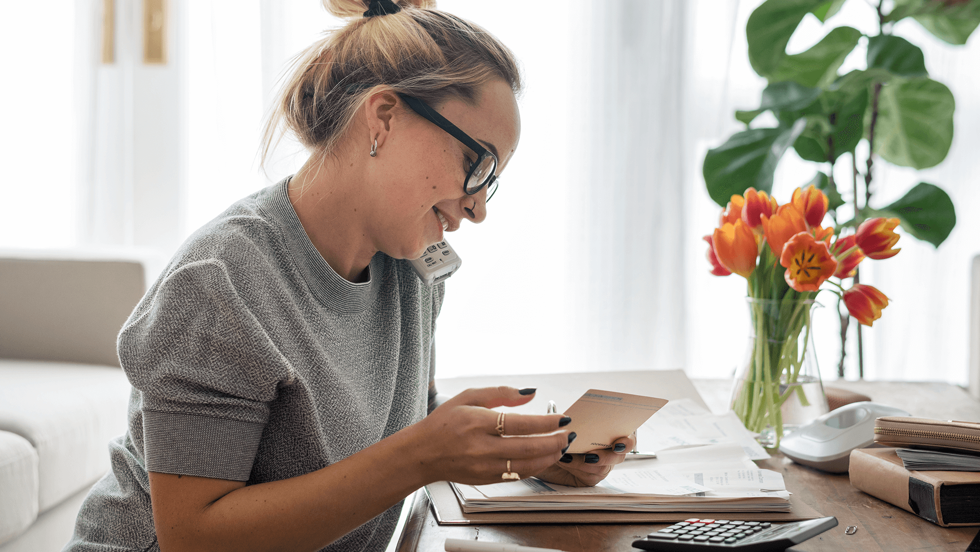A focused individual multitasking at a home office, conversing on the phone while jotting down notes, with fresh flowers adding a touch of vibrancy to the workspace.