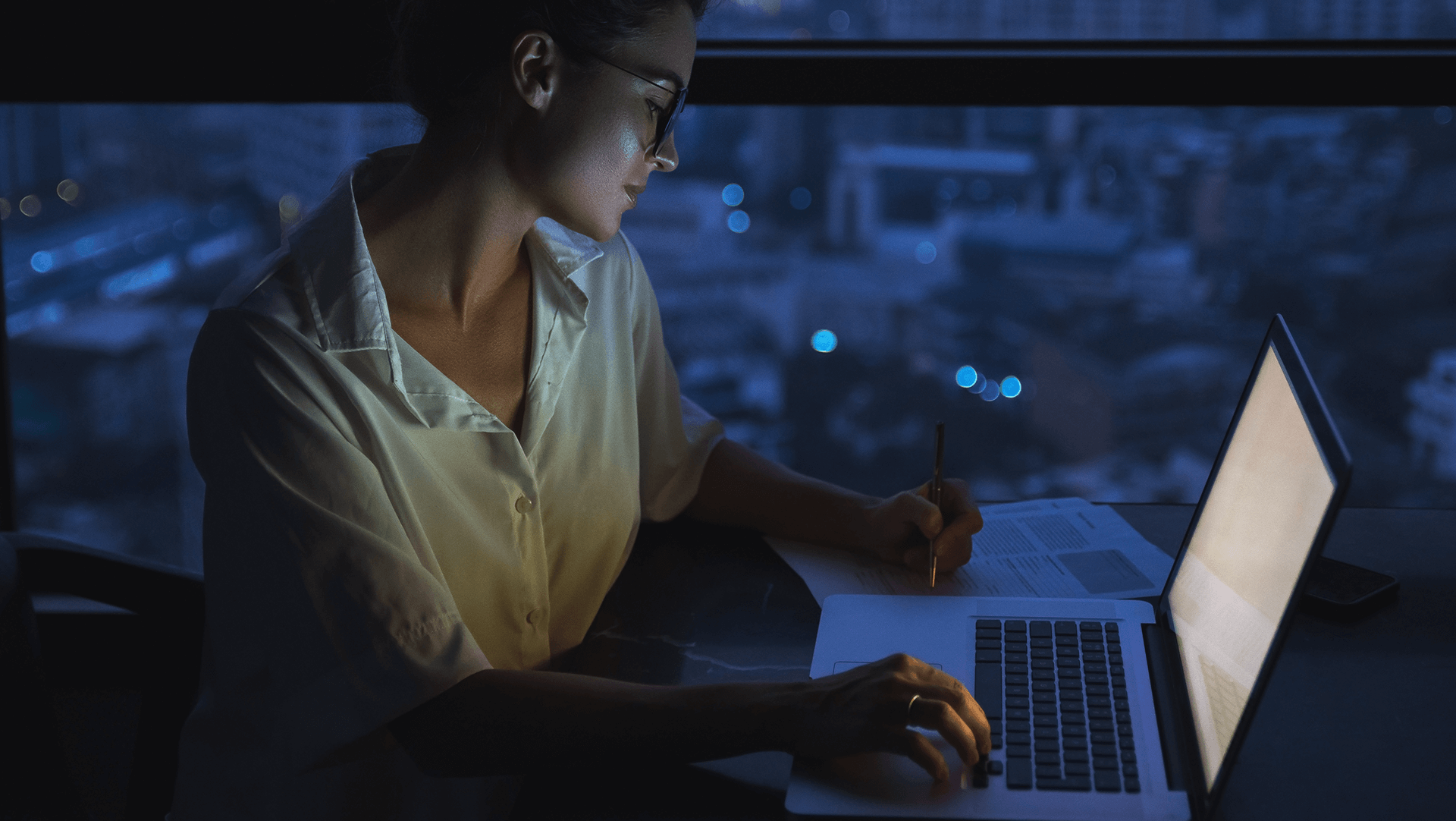 A focused individual working late at night on a laptop with papers, illuminated by the screen's glow against a backdrop of city lights.