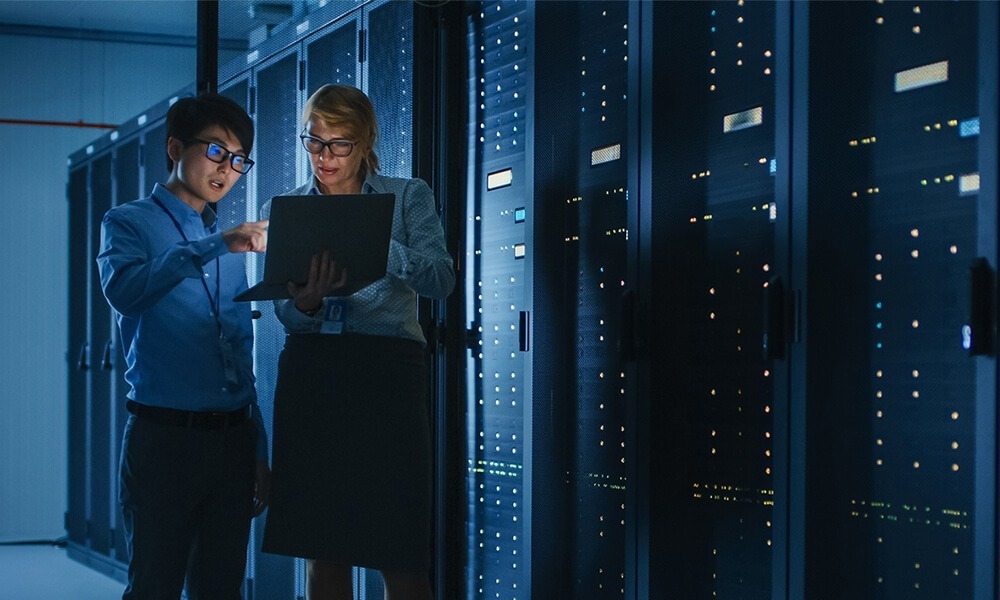 Two it professionals analyzing data on a laptop in a server room at night.