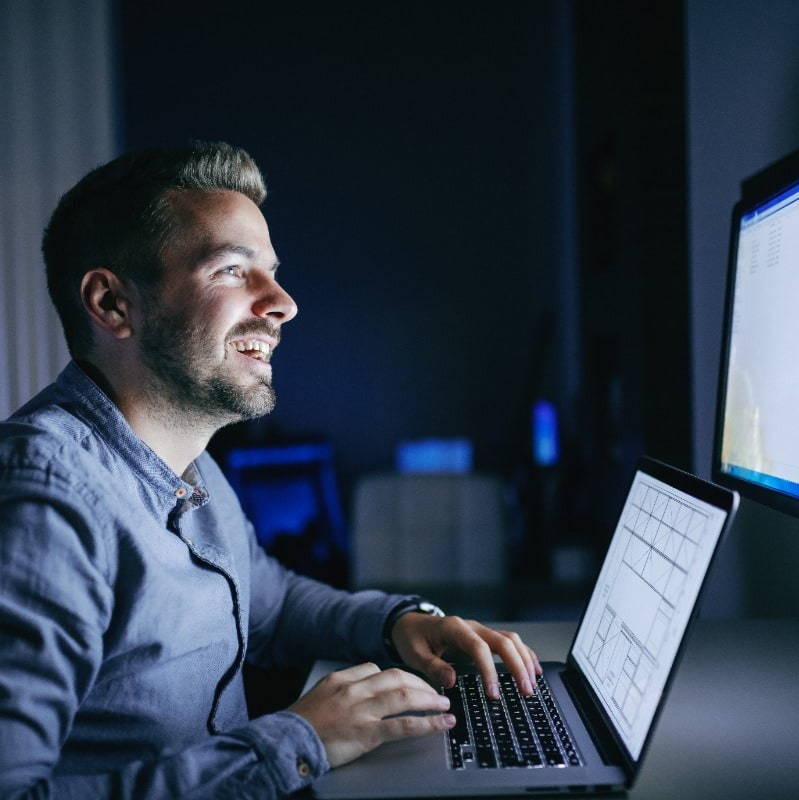 a middle aged man working infront of his laptop in a dim lit room while smiling