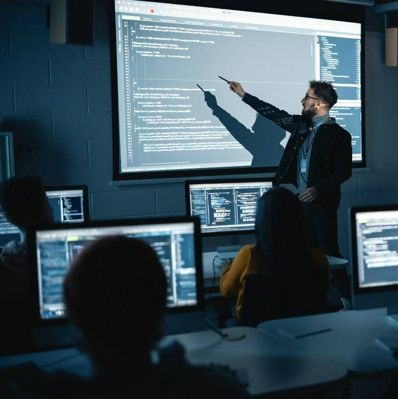 Instructor explaining code on a large screen to students in a modern computer lab.