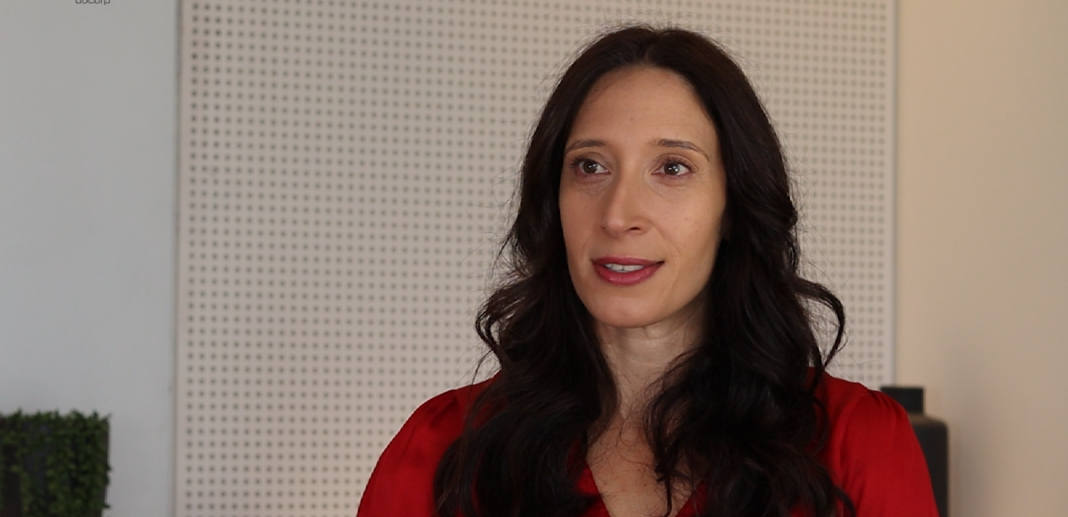 A professional woman in a red blouse speaking during an office meeting, with a modern, minimalist background featuring a perforated wall panel.