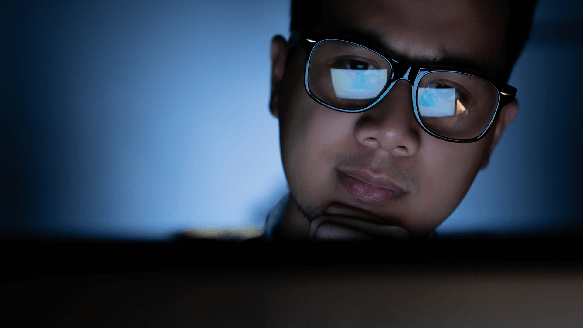 A focused individual wearing glasses is illuminated by the glow of a computer screen in a dimly lit room.