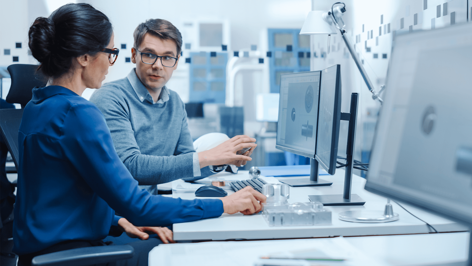 Two professionals discussing work at a computer station in a modern office environment.