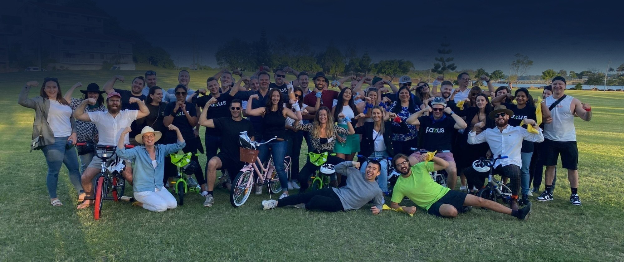 A large, cheerful group of people posing together outdoors on a grassy field, some sitting and some standing, with several individuals playfully holding up bicycles.