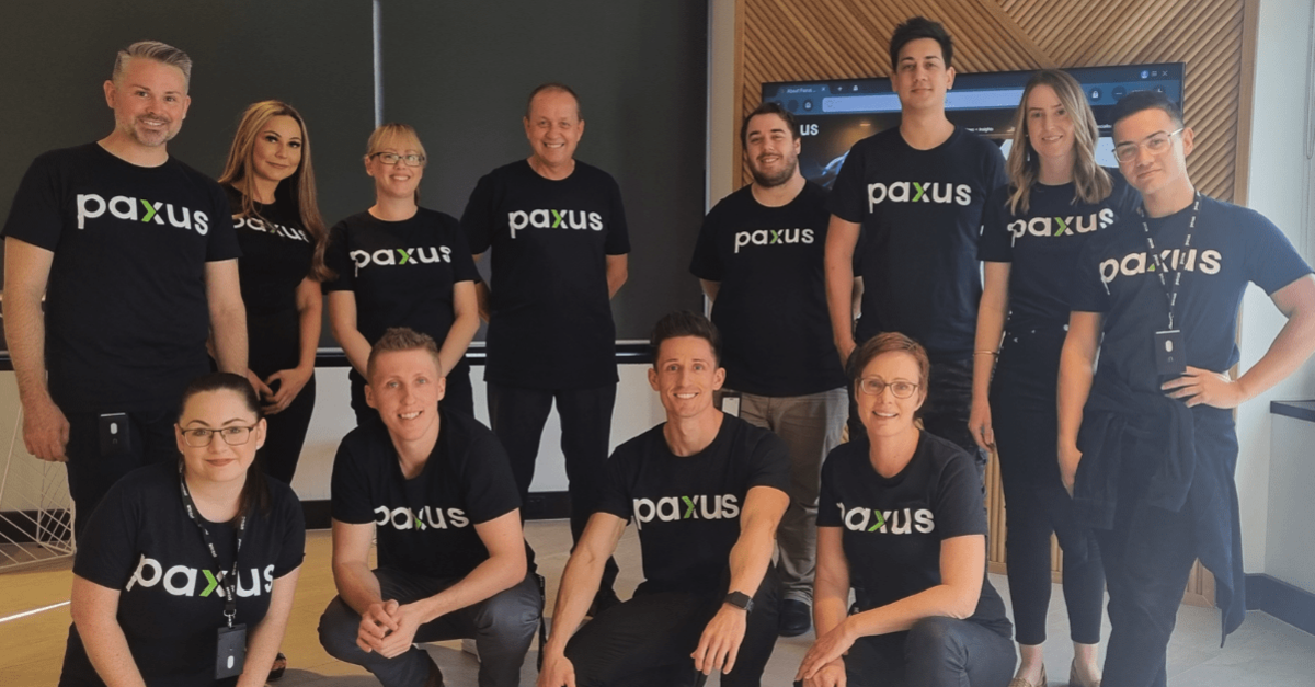 Team members proudly wearing Paxus-branded shirts, smiling cheerfully towards the camera