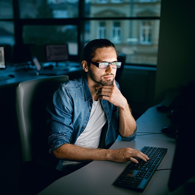 A focused individual wearing glasses and a denim shirt, deep in thought while working at a computer in a dimly lit office environment.