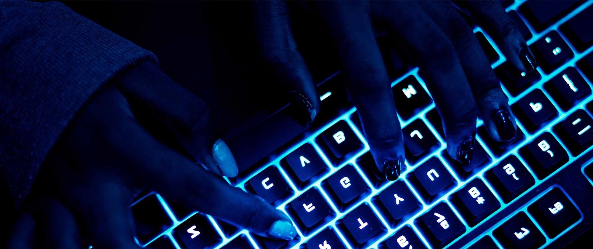 Hands typing on a backlit keyboard in a dimly lit environment, highlighting the interplay of technology and user interaction.